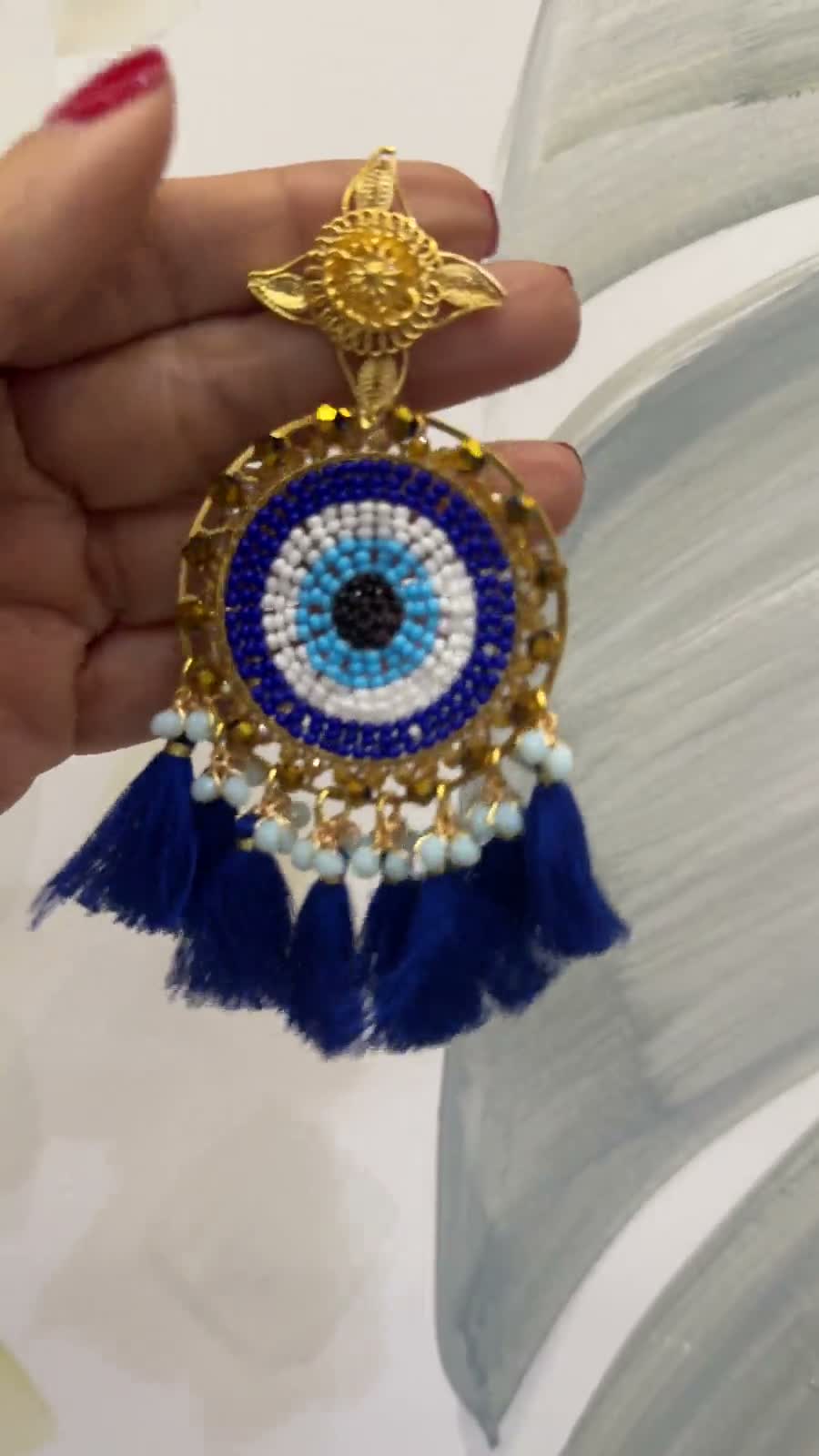 Arete circular con un ojo tejido en mostacilla azul, blanca y dorada con borlas